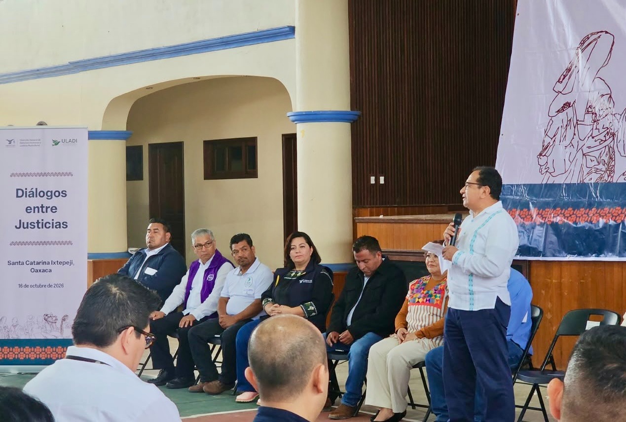 Ponente de pie con micrófono ante una mesa de participantes en el encuentro Diálogos entre Justicias en Santa Catarina Ixtepeji, Oaxaca