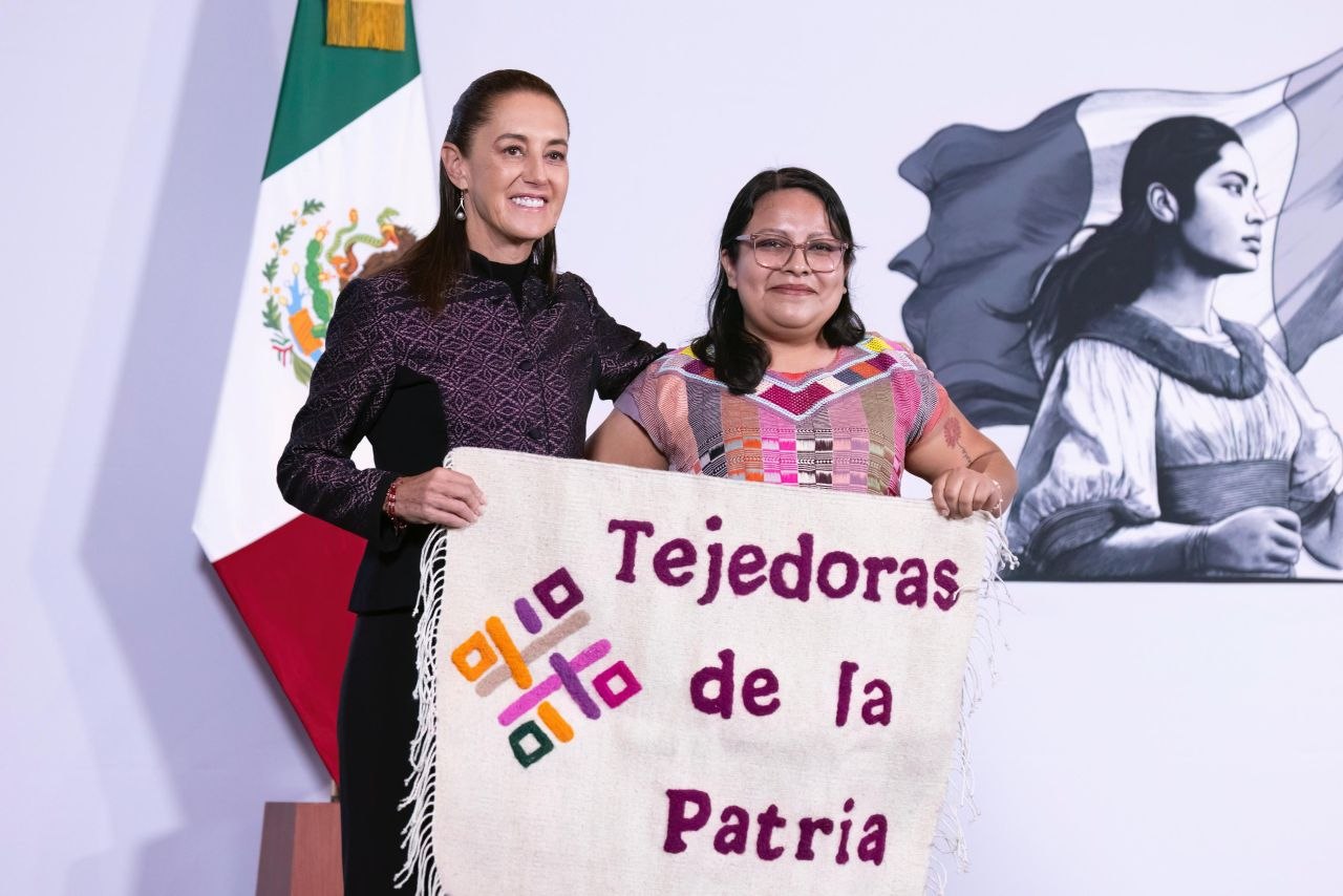 Dos mujeres sostienen un textil con la frase “Tejedoras de la Patria” frente a una bandera de México y una ilustración