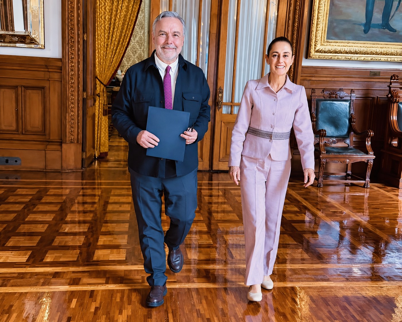 Dos personas caminan en un salón de Palacio Nacional; una de ellas sostiene una carpeta azul