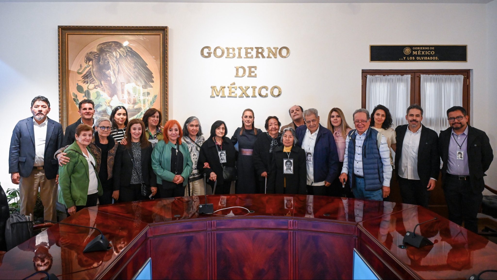 Grupo de personas posa en una sala oficial del Gobierno de México, algunas con fotografías colgadas al cuello