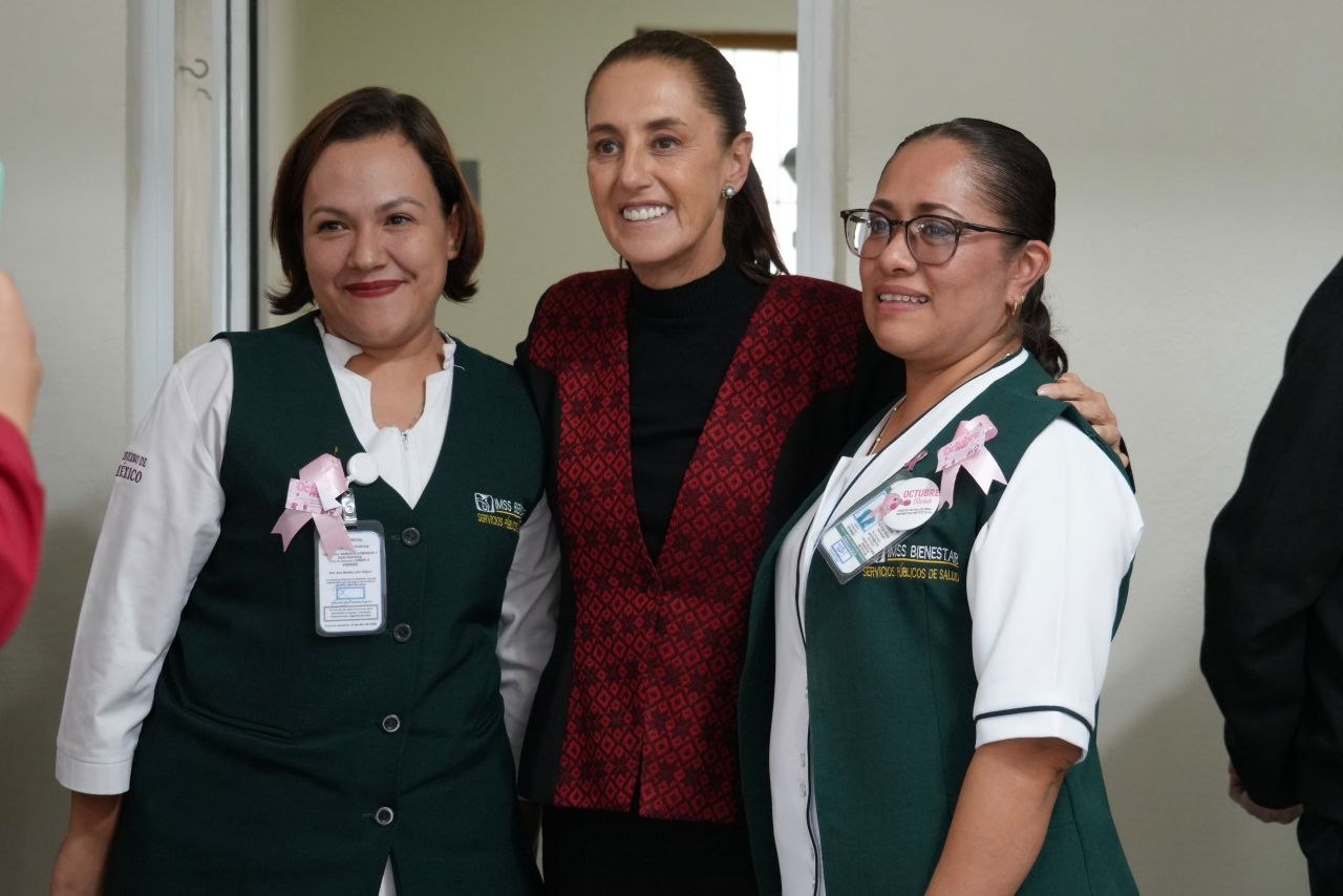 Tres mujeres posan para una fotografía; dos portan uniformes verdes con insignias de IMSS Bienestar y listones rosas.