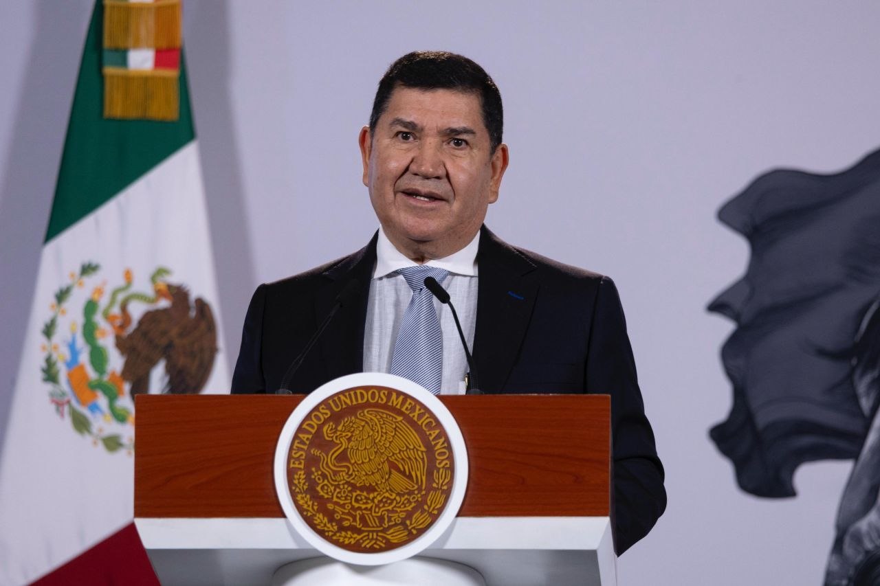 Un funcionario habla ante un podio con el escudo nacional y la bandera de México durante una conferencia de prensa.