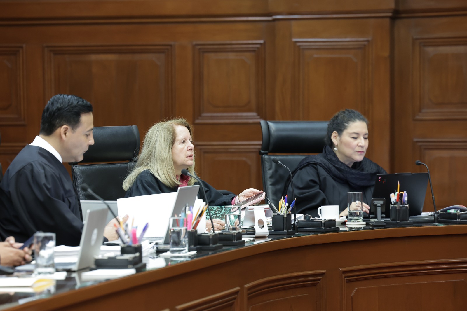 Tres personas con vestimenta judicial participan en una sesión de tribunal frente a computadoras y micrófonos